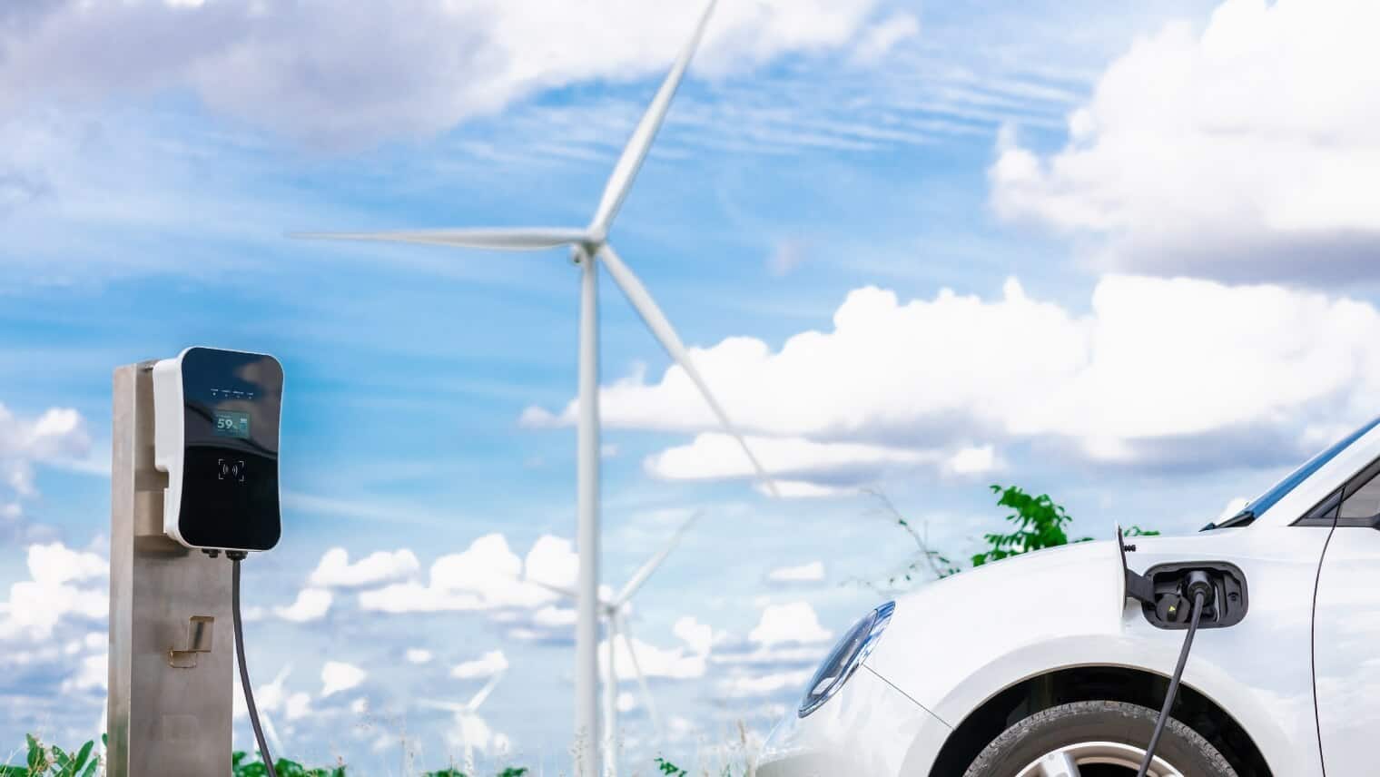 Coche eléctrico enchufado a una estación de carga inteligente con una moderna turbina eólica al fondo, que representa la energía limpia habilitada por IoT.